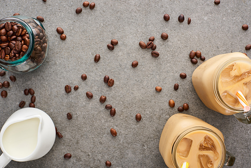 top view of ice coffee in glass jars with straws near milk and coffee grains on grey background