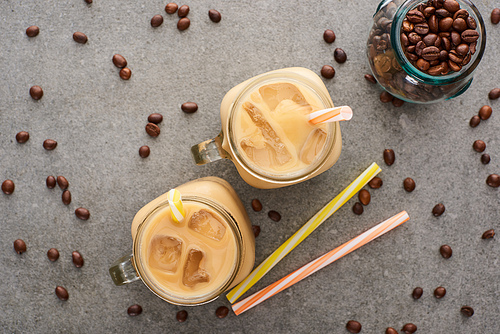 top view of ice coffee in glass jars near straws and coffee grains on grey background