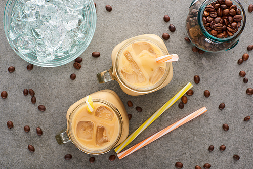 top view of ice coffee in glass jars near straws, coffee grains and ice cubes on grey background