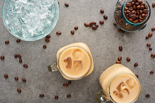 top view of ice coffee in glass jars near coffee grains and ice cubes on grey background