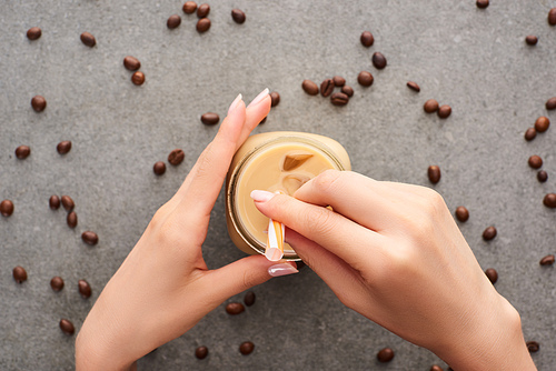 partial view of woman holding glass jar with straw and ice coffee near coffee grains on grey background