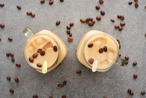 top view of ice coffee in glass jars with straws and coffee grains on grey background