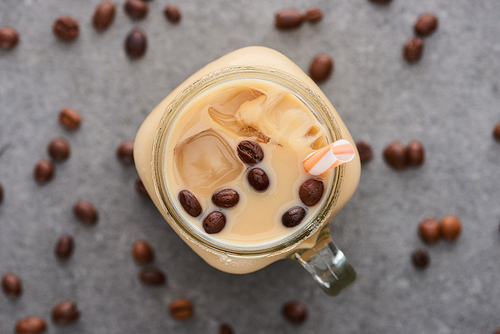 selective focus of ice coffee in glass jar with straw near coffee grains on grey background