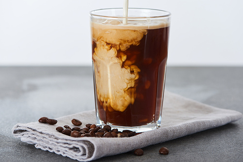 milk pouring into ice coffee in glass on napkin with coffee grains isolated on white