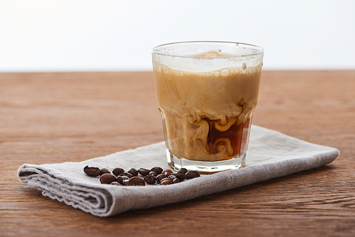 ice coffee mixing with milk in glass on napkin with coffee grains on wooden table isolated on white