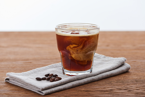 ice coffee mixing with milk in glass on napkin with fesh coffee grains on wooden table isolated on white