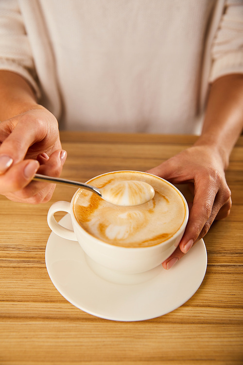 selective focus of woman holding spoon with cappuccino foam near wooden table