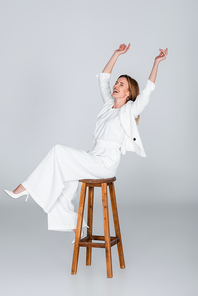 full length of excited woman in white outfit sitting on stool on grey