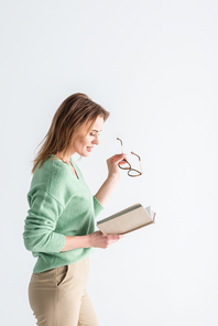 cheerful woman holding glasses and reading book isolated on white