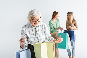 surprised senior woman looking at shopping bag near daughter and granddaughter on blurred background
