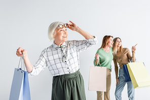 happy senior woman with shopping bag adjusting glasses near daughter and granddaughter on blurred background