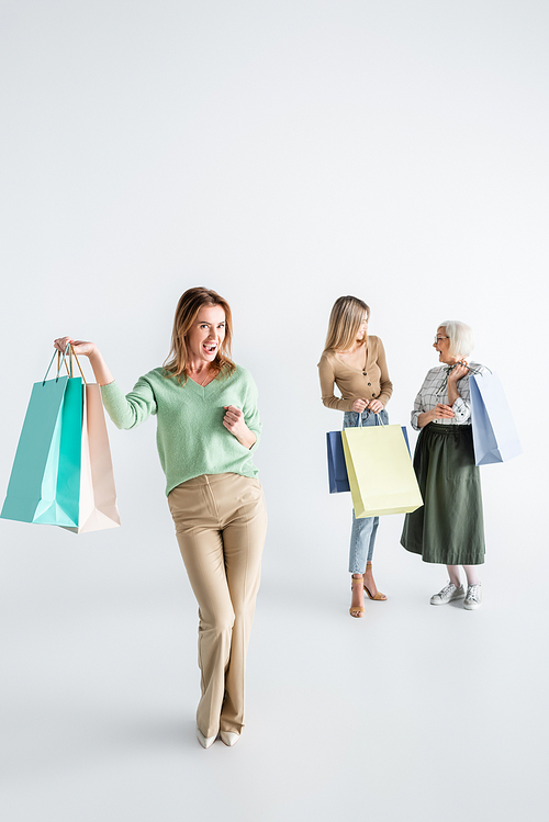 full length of happy woman with shopping bags near daughter and senior mother on blurred white background