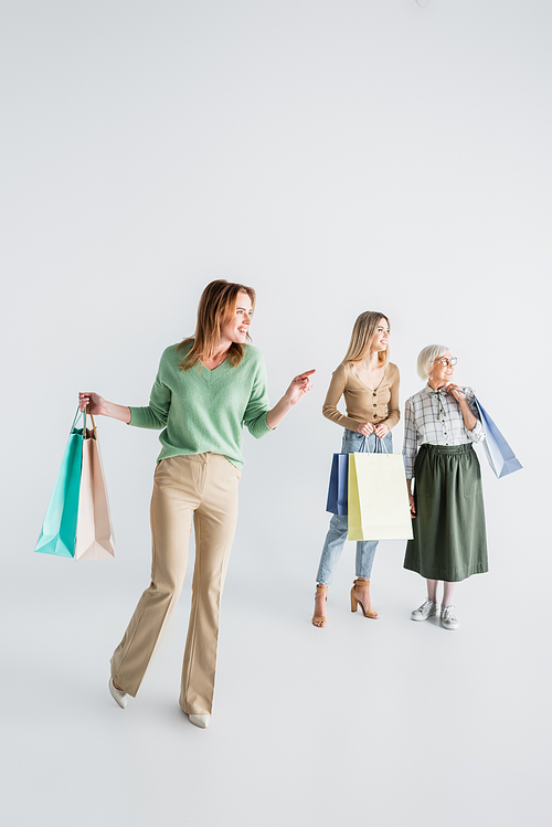 full length of happy woman holding shopping bags and pointing with finger near daughter and senior mother on blurred white background