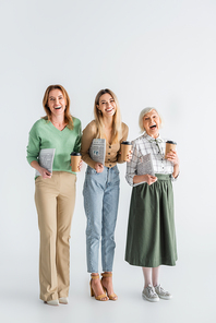 full length of three generation of cheerful women holding newspapers and paper cups on white