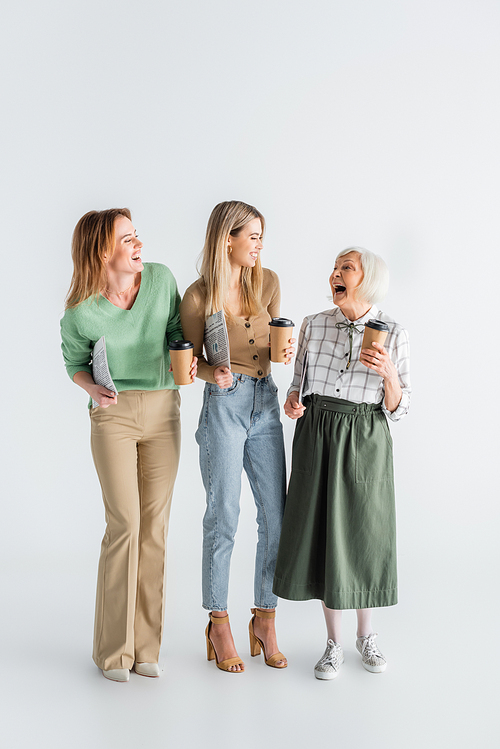 full length of three generation of smiling women holding newspapers and paper cups on white