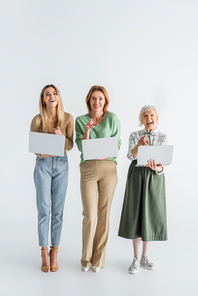 full length of three generation of cheerful women pointing with fingers and holding laptops on white