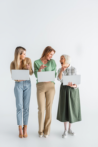full length of three generation of cheerful women pointing with fingers, looking at each other and holding laptops on white