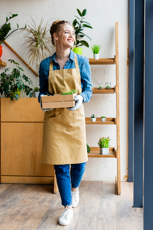 happy woman in gloves holding wooden box with green plants and looking away