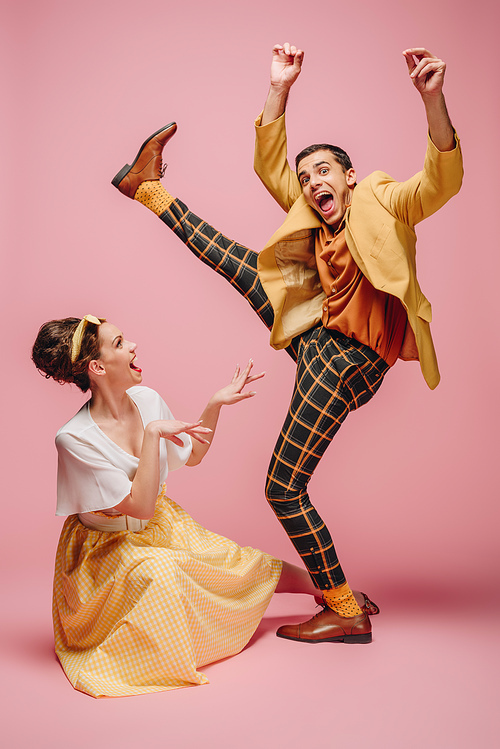 cheerful girl sitting on floor and excited man raising leg while dancing boogie-woogie on pink background