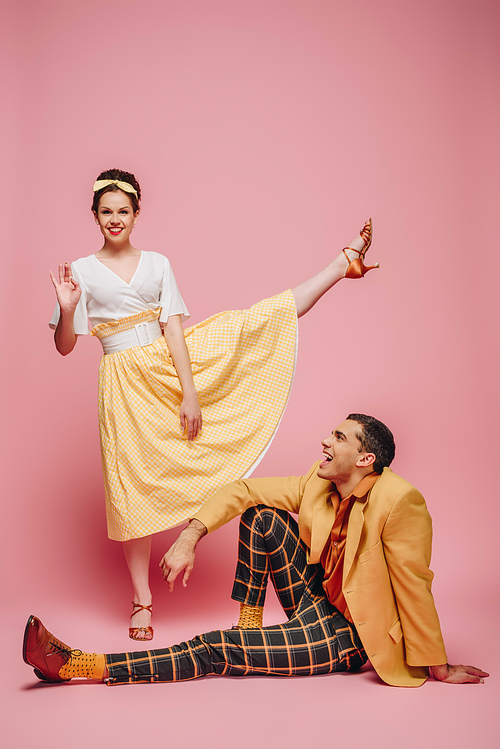 cheerful man sitting on floor and girl waving at camera while dancing boogie-woogie on pink background