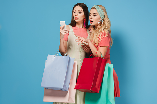 surprised brunette and blonde women in dresses holding shopping bags and using smartphone