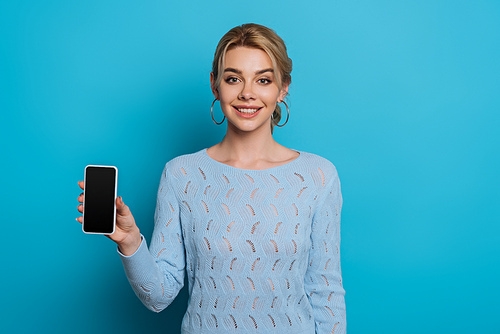 cheerful girl smiling at camera while showing smartphone with blank screen on blue background