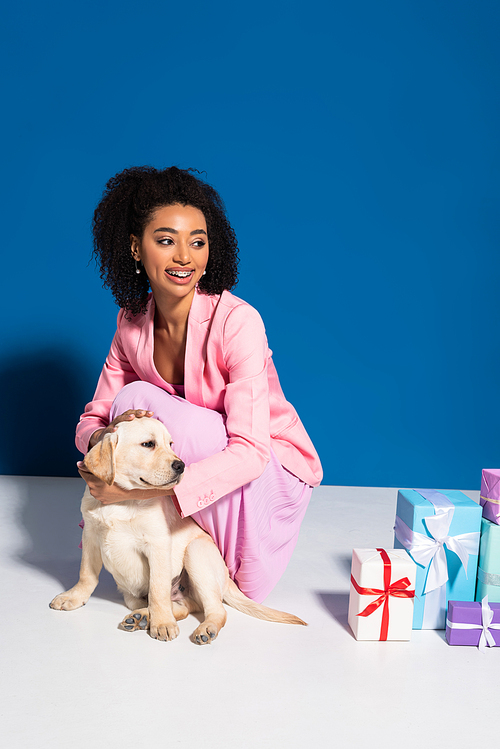 smiling african american woman with golden retriever puppy near gifts on blue background