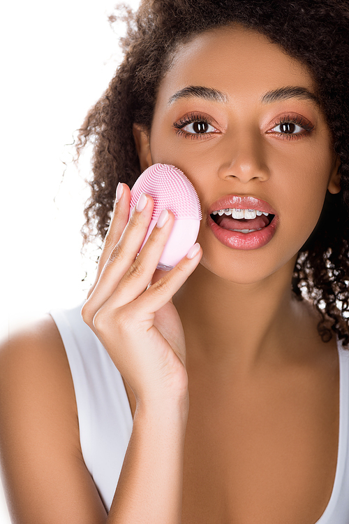 surprised african american girl with braces using silicone cleansing facial brush, isolated on white
