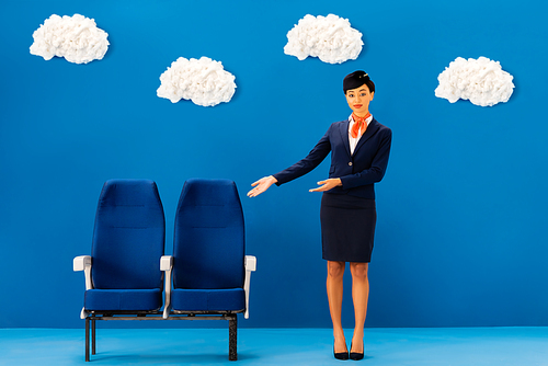 african american flight attendant pointing with hands at seats on blue background with clouds