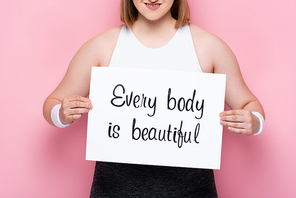 cropped view of smiling overweight girl holding placard with every body is beautiful inscription on pink