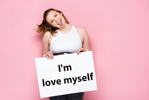 cheerful overweight girl holding placard with i love myself inscription and sticking out tongue on pink