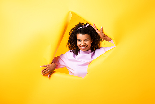 happy african american woman  through hole in ripped paper on yellow background