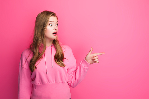 shocked beautiful woman in hoodie pointing aside on pink background