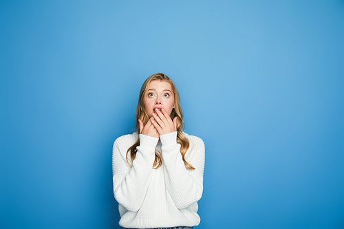 shocked blonde beautiful woman in sweater covering mouth isolated on blue
