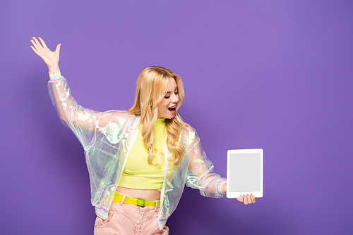 happy blonde young woman in colorful outfit presenting digital tablet on purple background