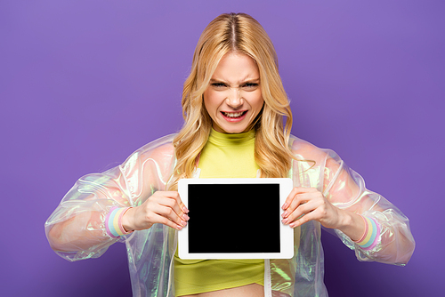 confused blonde young woman in colorful outfit presenting digital tablet on purple background