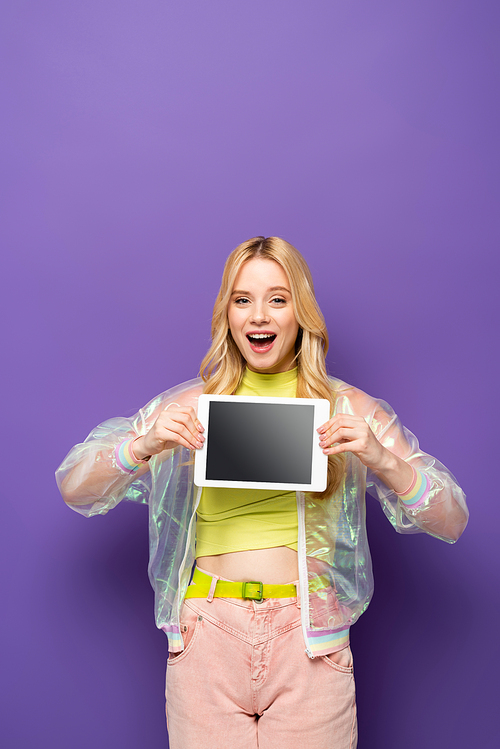 happy blonde young woman in colorful outfit holding digital tablet on purple background