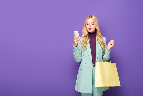 blonde young woman in fashionable turquoise blazer with smartphone and shopping bag on purple background