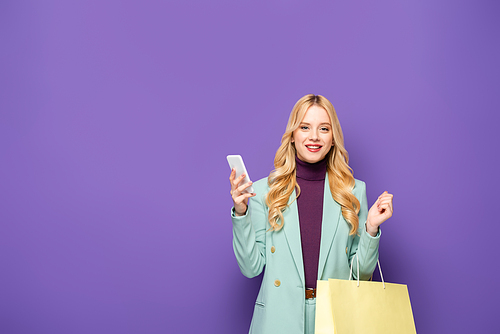 smiling blonde young woman in fashionable turquoise blazer with smartphone and shopping bag on purple background