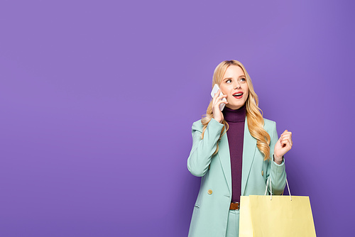 blonde young woman in fashionable turquoise blazer talking on smartphone and holding shopping bag on purple background
