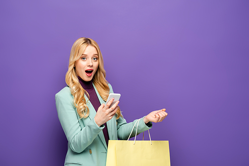 shocked blonde young woman in fashionable turquoise blazer with smartphone and shopping bag on purple background