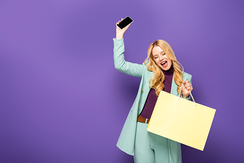 happy blonde young woman in fashionable turquoise blazer with smartphone and shopping bag on purple background