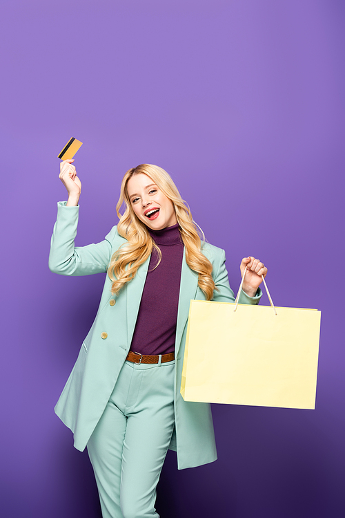 happy blonde young woman in fashionable turquoise blazer with credit card and shopping bag on purple background