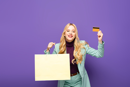 happy blonde young woman in fashionable turquoise blazer with credit card and shopping bag on purple background