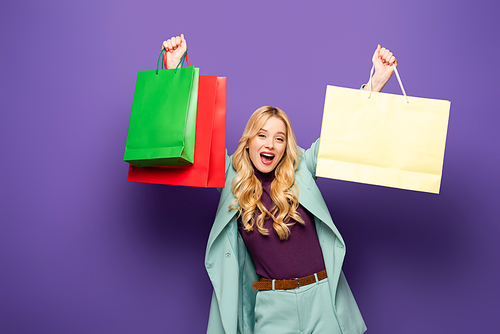 happy blonde young woman in fashionable turquoise blazer with shopping bags on purple background