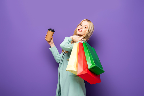 happy blonde young woman in fashionable turquoise blazer with shopping bags and paper cup on purple background