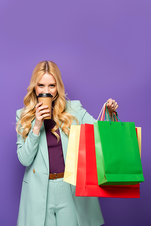 blonde young woman in fashionable turquoise blazer with shopping bags and paper cup on purple background