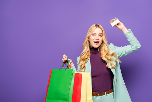happy blonde young woman in fashionable turquoise blazer with shopping bags and credit card on purple background