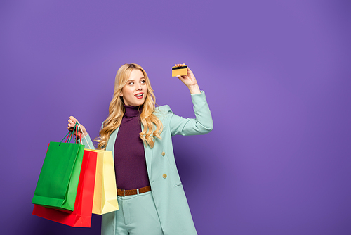blonde young woman in fashionable turquoise blazer with shopping bags and credit card on purple background