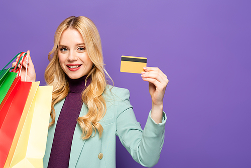 smiling blonde young woman in fashionable turquoise blazer with shopping bags and credit card on purple background